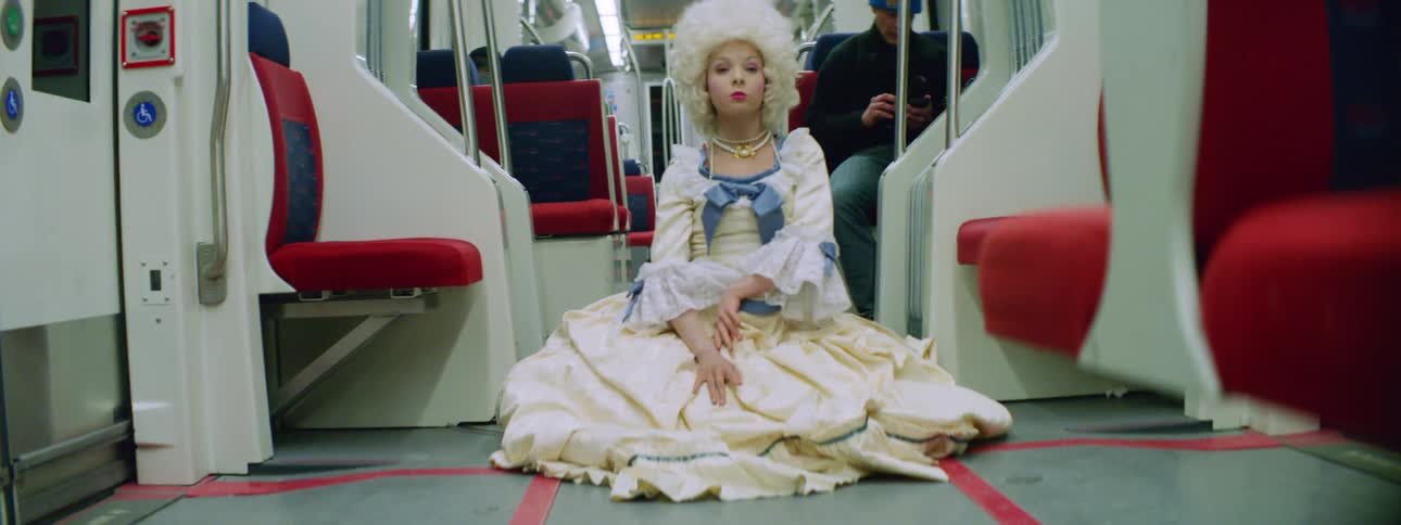 A young woman in an old fashioned dress and wig dances while sitting on the floor of a subway train