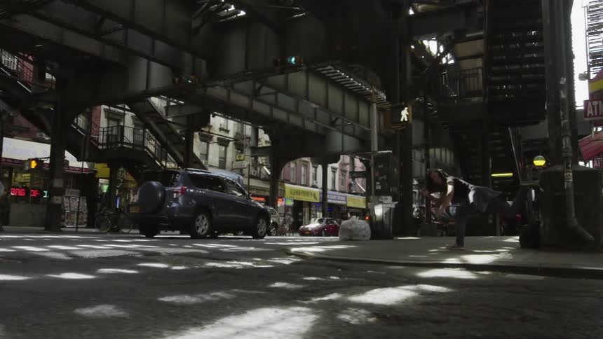 A woman is dancing on the corner under a train station as cars and people walk by