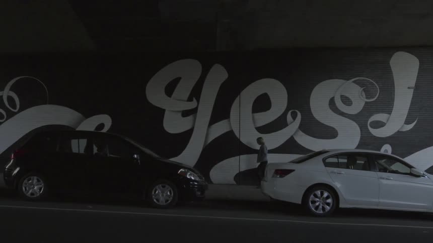 A person walks slowly under a bridge next to parked cars