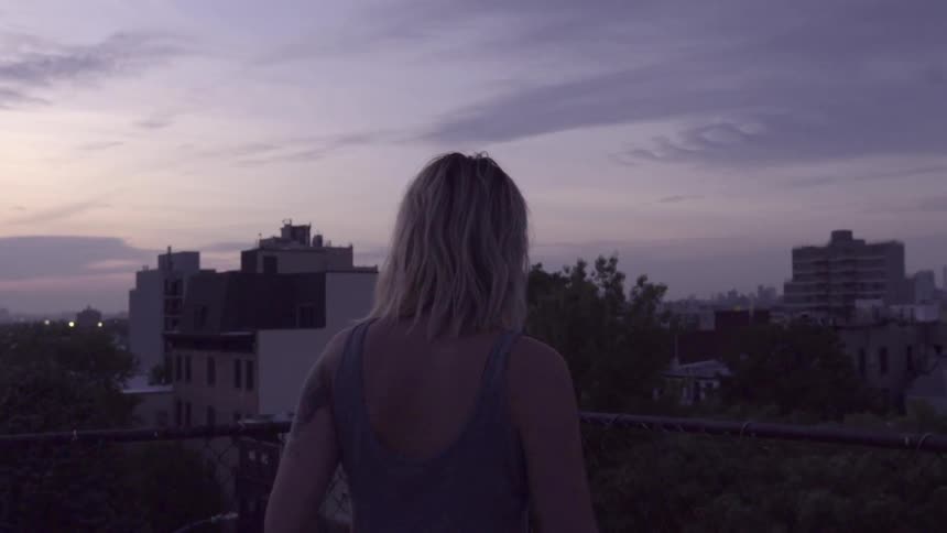 A woman walks over to a fence on a rooftop at sunset