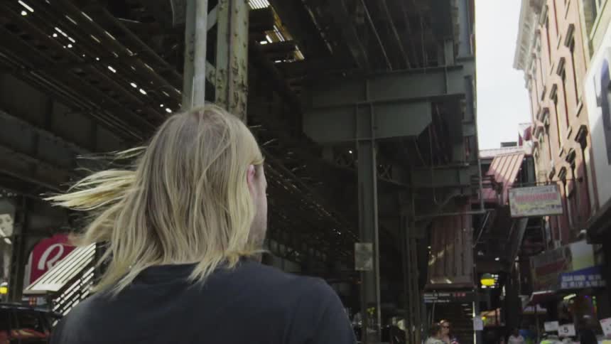 A man with long blonde hair walks in a city on a sunny day