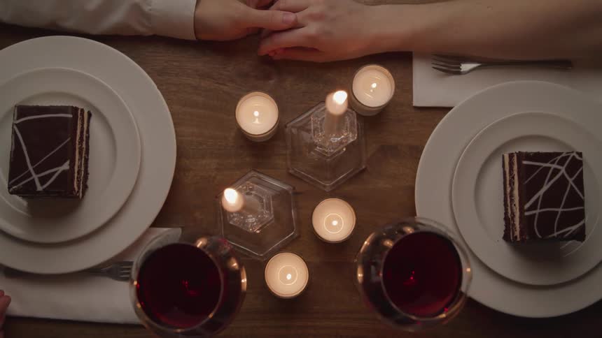 A man and a woman sitting at a table touching hands with candles wine and dessert