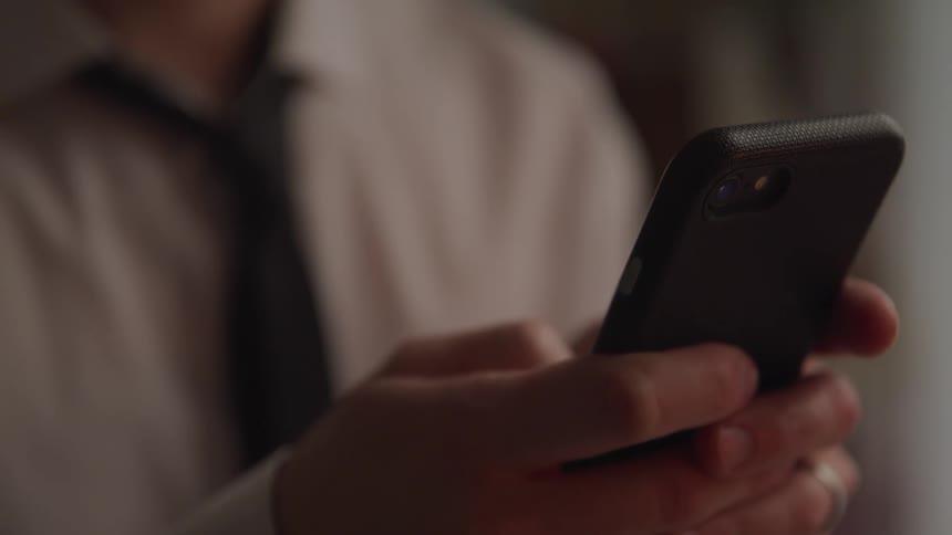 Male wearing a tie holds a smart phone with two hands while using his thumb to scroll down on the screen
