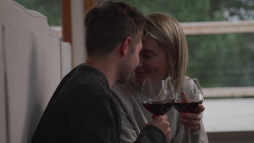 A man and woman in a home each hold a glass of wine as they rest their heads together