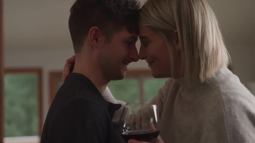 A woman holding a glass of wine smiles as her and her boyfriend put their heads together