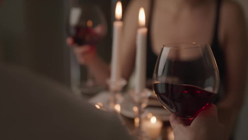 A man and woman swirl wine in their wine glass as they face each other across a dining table cover in candles