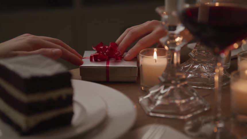 A man slides a small gift across a dinner table to the woman sitting on the other side