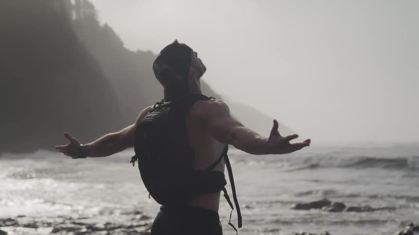 Shirtless young man wearing a backpack with arms wide open at the beach