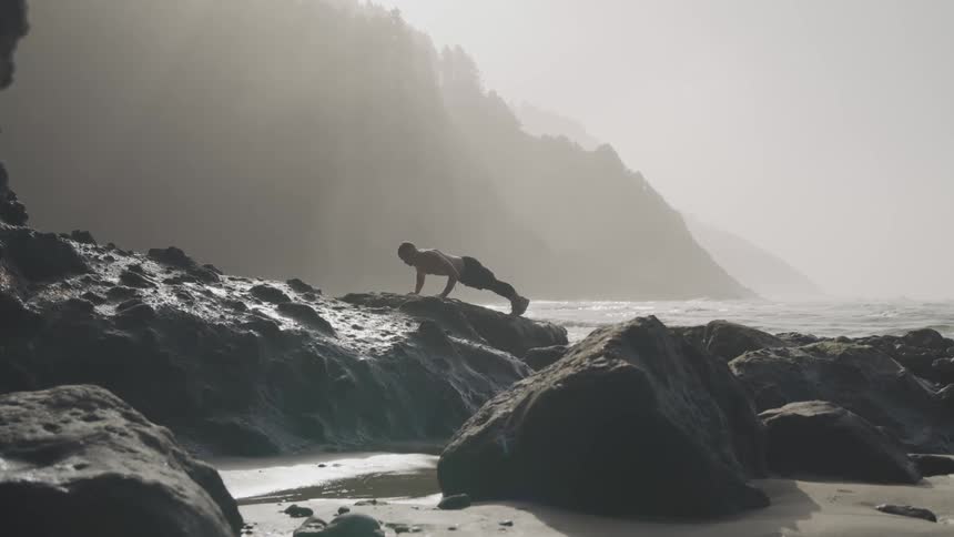 Young shirtless fit man does push ups over the big rocks on the coast of a beach next to the mountains