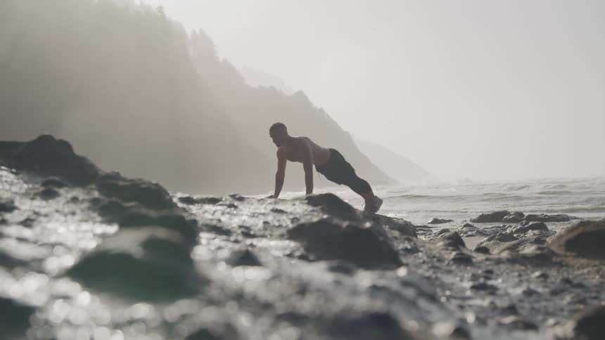 A shirtless man does pushups on a rock formation next to the sea on a foggy morning