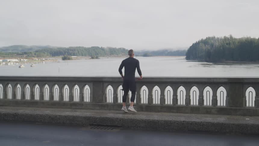 A man standing on a bridge jumps as he looks at the water and a car drives past