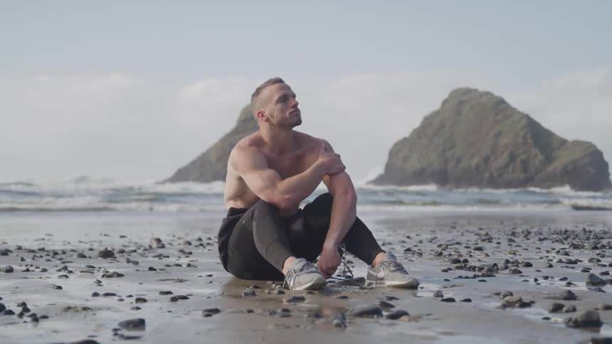 A muscular man sits with his shirt off on the beach