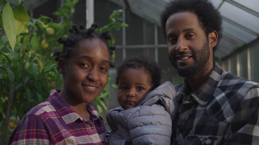 A man and woman hold a child between them as they smile into the camera while standing in a greenhouse at night