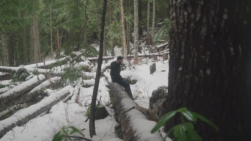 A man sits on a fallen down tree trunk in a snowy forest