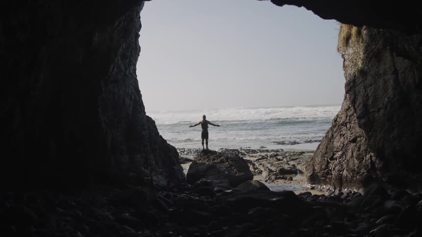 Man stands on large boulder in mouth of cave facing ocean
