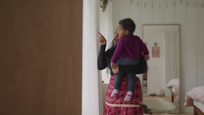 A woman holds a little girl as she stands near a window and points outside