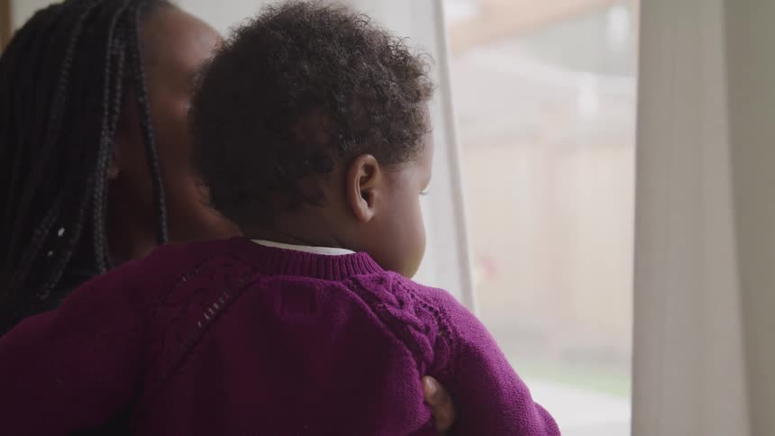 A woman holds her infant son next to a window looking outside