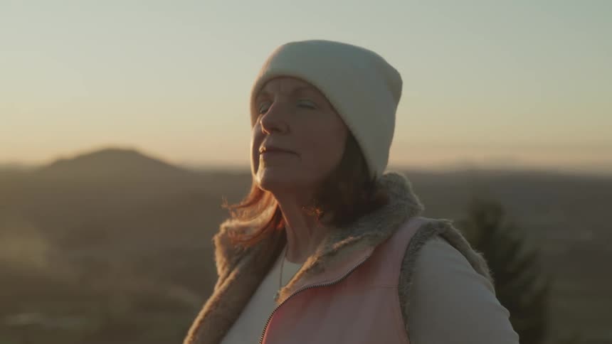 A woman is outdoors wearing a hat while the sun is setting