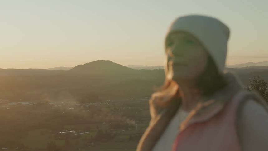 A woman wearing warm clothes and a beanie stands with a mountain range in the distance behind her