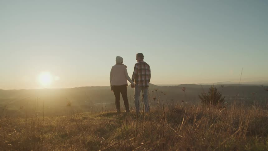 Man wearing a plaid shirt and woman wearing a pink coat and a white beanie hold hands and look at each other on the edge a cliff at sunset