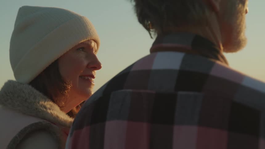 Woman with a white beanie looks at man with a tartan shirt watching sunset