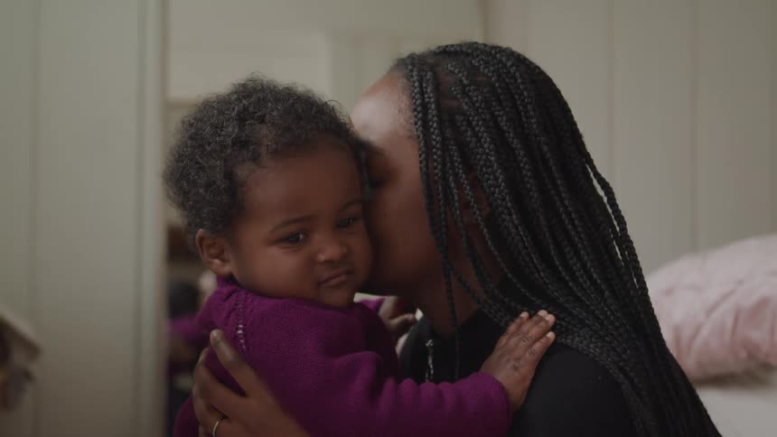 A woman with long braids hugs her infant son as they sit on the floor in their bedroom