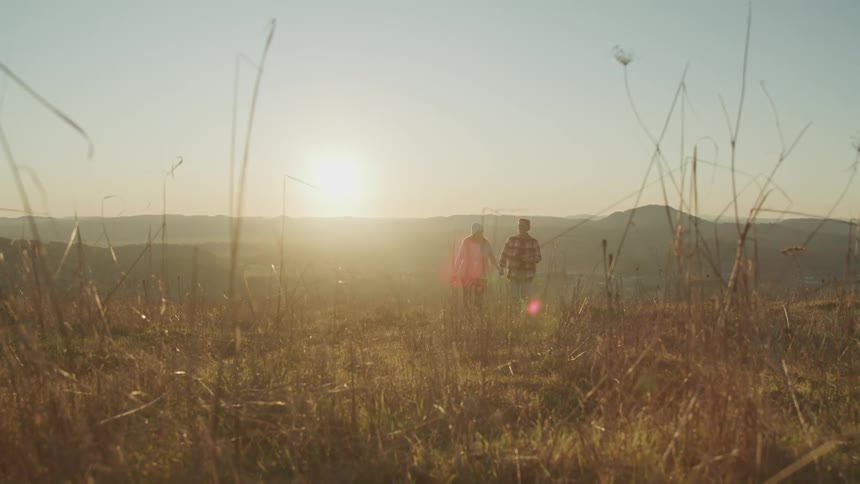 A man and woman hold hands and laugh as they walk through a field turning brown in fall
