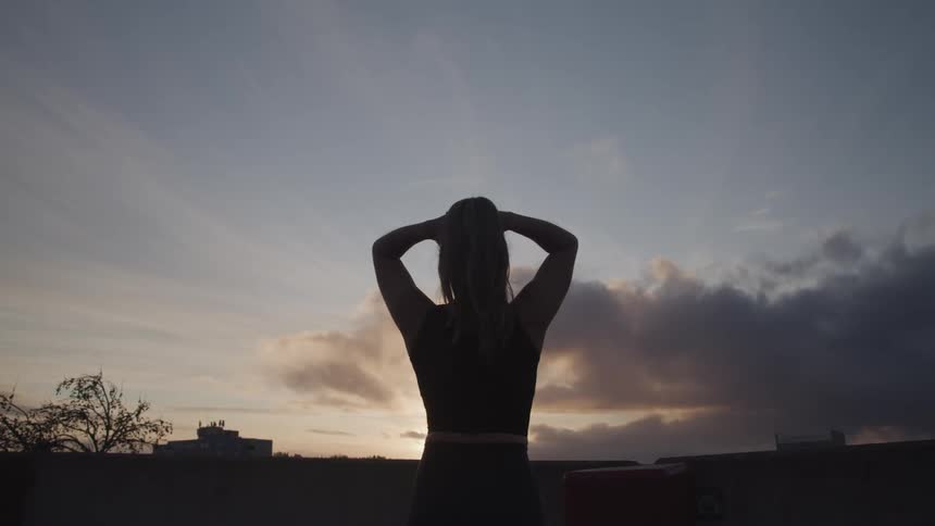 Woman stands looking toward red tinted horizon with hands against her head