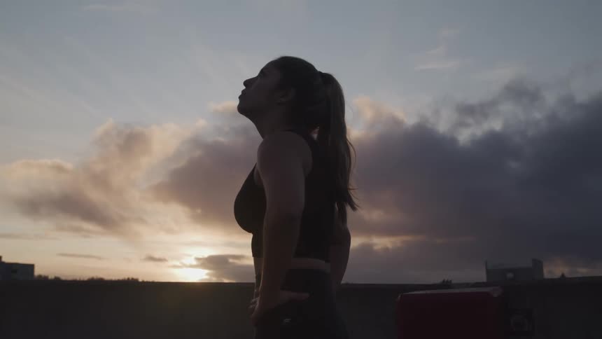 A woman in athletic wear stands on the rooftop of a building in a city and gazes at the sky