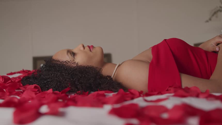 A woman with a red dress and a necklace laying on a bed with red flower petals