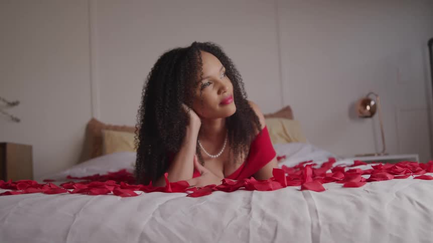 A woman with a red dress and a necklace laying on a bed with red flower petals