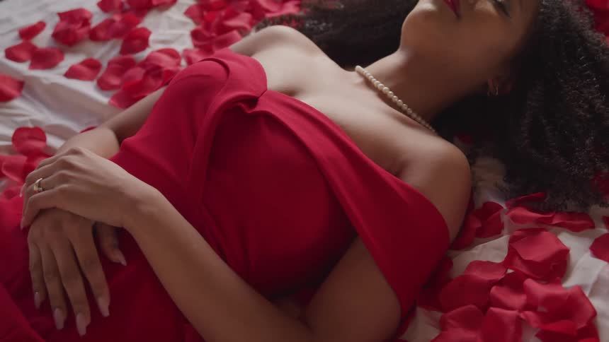 Young woman in a red dress and pearl necklace laying in bed surrounded with rose petals