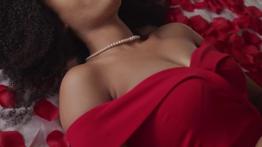Young woman in a red dress and pearl necklace laying in bed over red rose petals