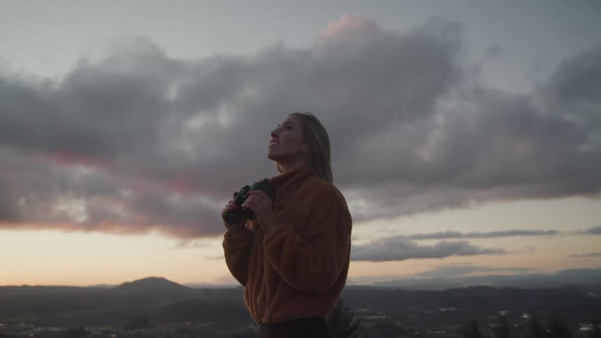 A woman with binoculars and a jacket looking towards the sky with clouds and the sky in the background