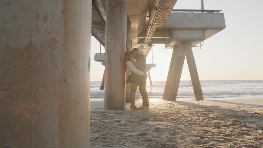 A couple embraces underneath a pier while standing on the sand