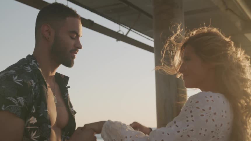 A man and a woman on a beach underneath a pier where he is kissing her hand