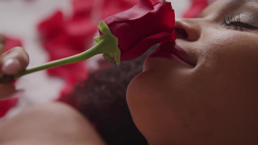 A woman lying on a bed covered in rose petals slowly brings a rose to her nose