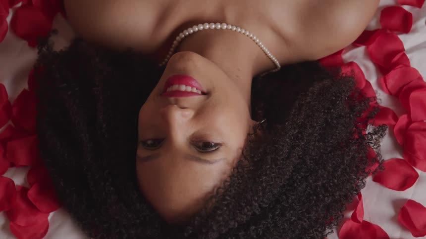 A smiling young woman lying on a bed covered in rose petals turns her head to gaze into the camera