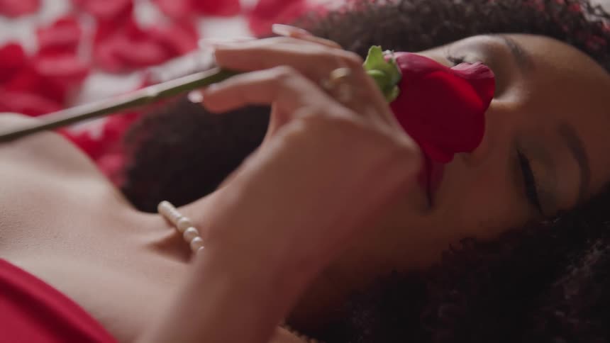 Woman wearing pearls lies on back among scattered rose petals smelling red rose
