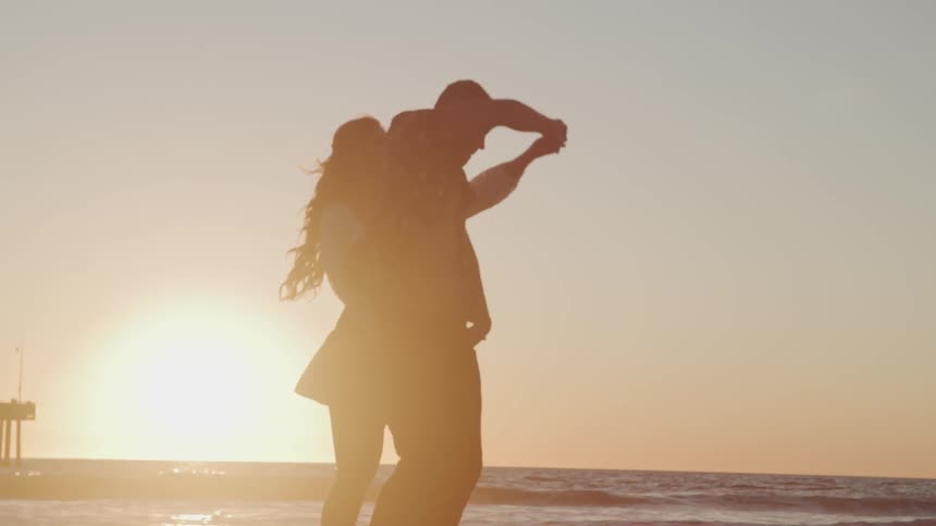 A man and woman dance on a beach in front of the setting sun