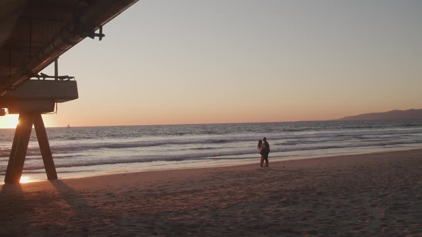 Young couple hugging on the sand beach at sunset next to a pier