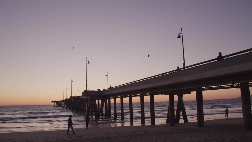 People walk across the beach and ocean side pier with a very low sun in the background
