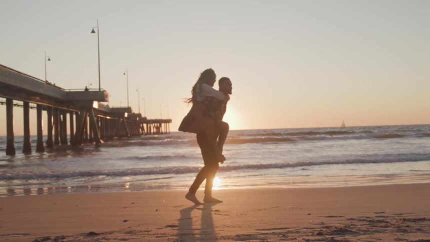 A man carries a woman on his back as he walks along a beach under a low sun