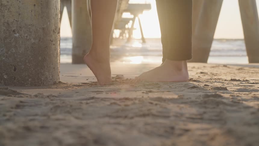 Woman stands on the tip of her toes to kiss a man under a boardwalk next to the ocean at sunset