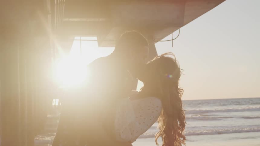A man and woman share a kiss under a pier on a beach on a sunny day