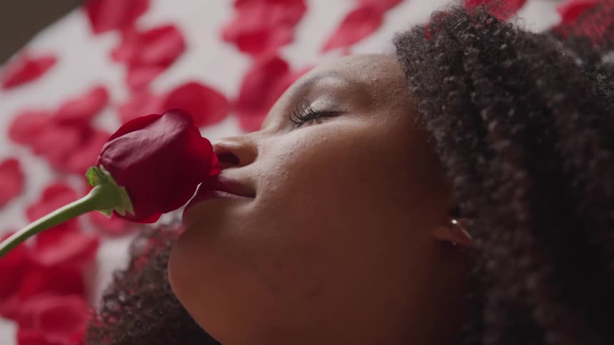 A woman lays on a bed covered in rose petals and takes a sniff of a single red rose