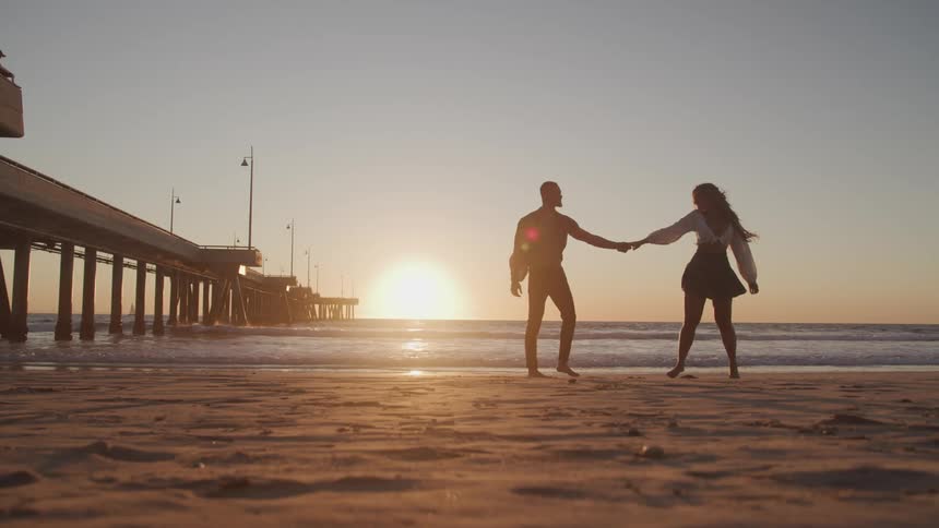 A man and woman dance together on a quiet beach in front of the low sun horizon