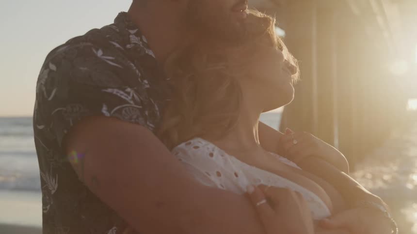 A man wraps his arms tightly around a woman as they stand under an ocean pier