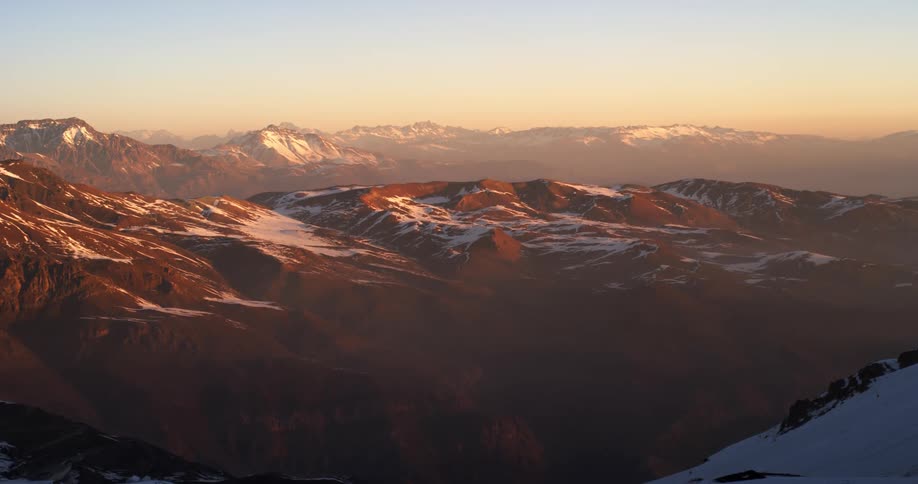 A range of desert mountains with patches of snow under an orange sky