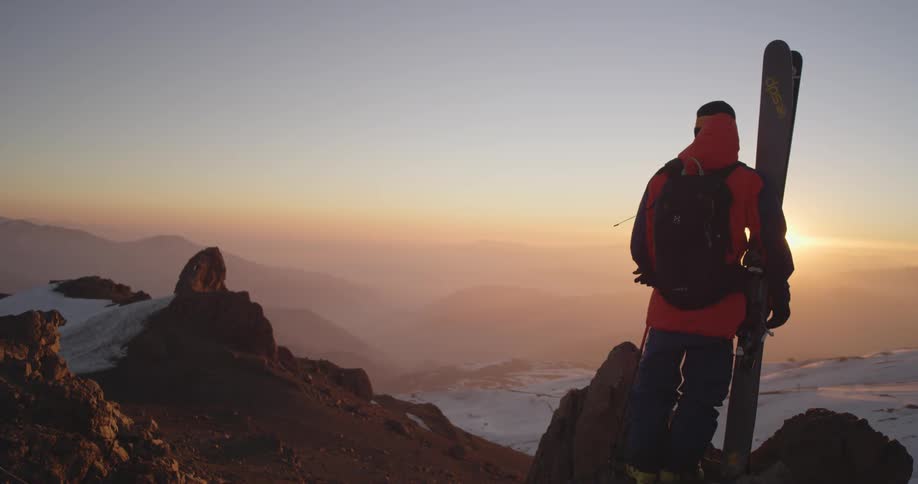 A man holding his skis looks out over a mountain at a sunset